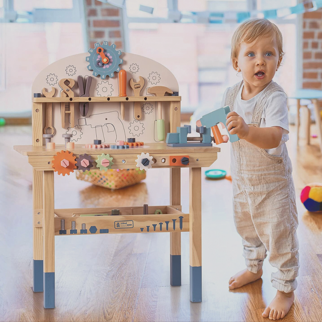 Child playing with a wooden toy workbench in a room with toys scattered around.